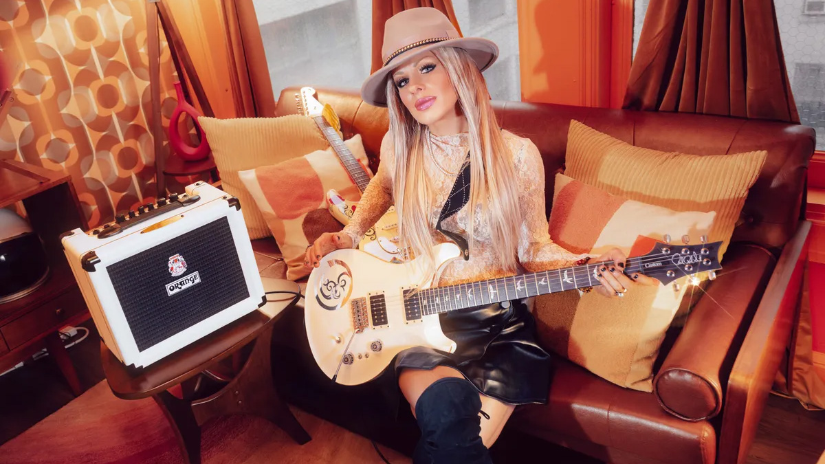 Orianthi with her PRS guitar, sitting on a sofa next to her signature Orange amp