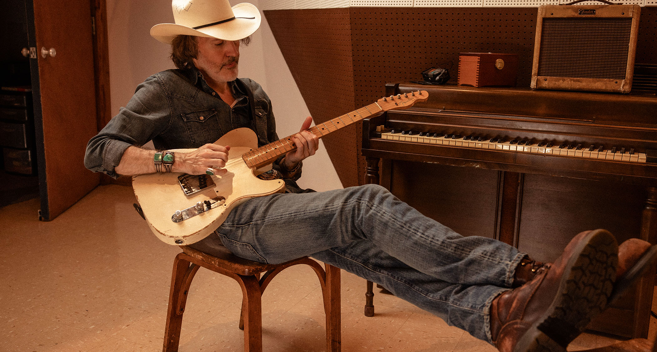 David Rawlings reclines on a chair in front of a piano and picks some notes on his Fender Telecaster. He wears a wide-brimmed hat.