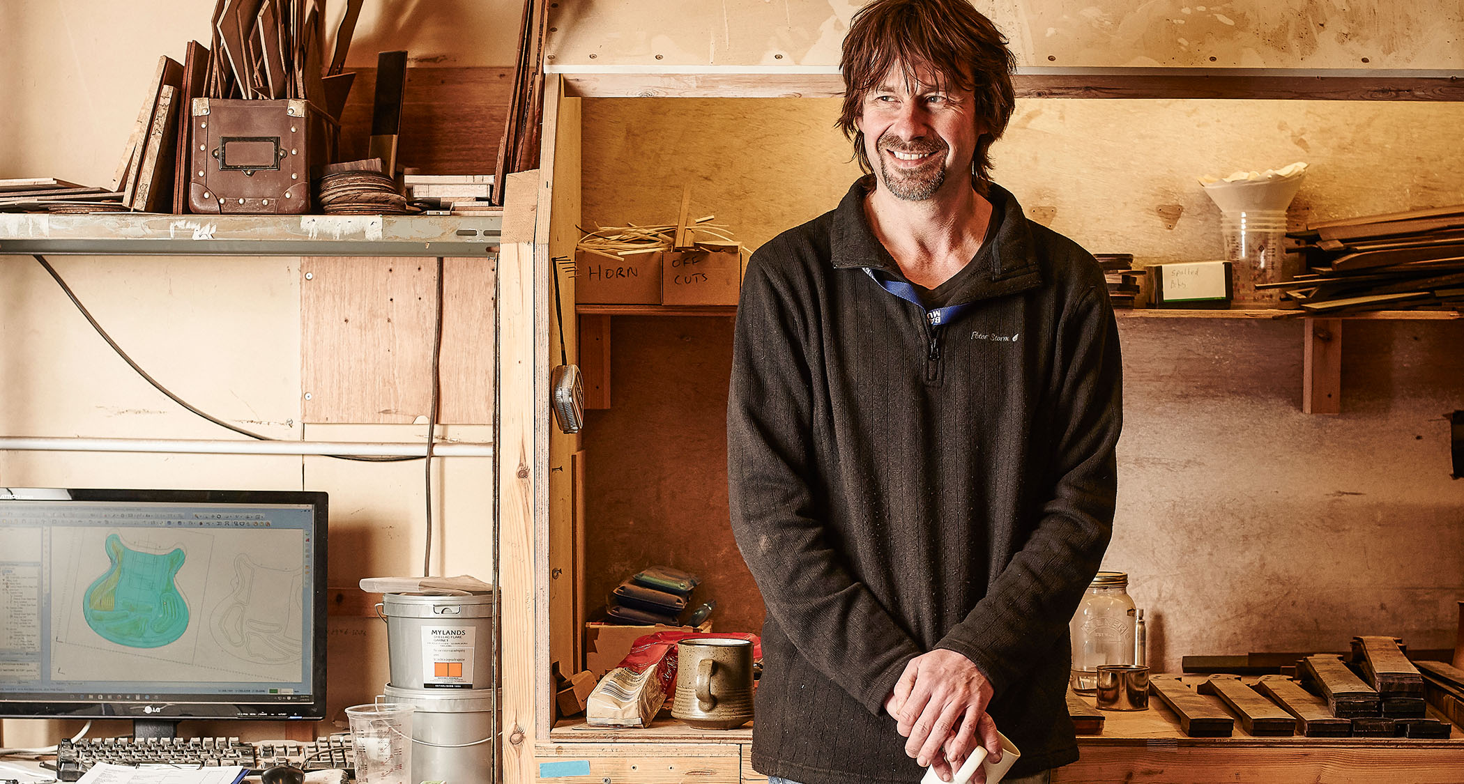 Esteemed British luthier Patrick James Eggle pictured in his workshop. He holds a mug in front of guitar parts, while a computer screen displays his work in progress.