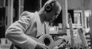 A black-and-white image of Eric Bibb laying down some fingerstyle acoustic guitar in the studio. He wears a suit jacket and monitoring headphones.