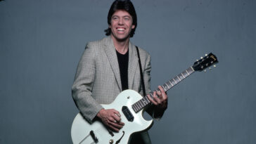 George Thorogood is shown in a studio portrait, wearing a tan jacket over a baclk t-shirt and pants. He is playing a guitar he has strapped on