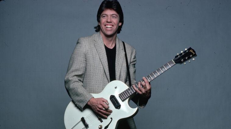 George Thorogood is shown in a studio portrait, wearing a tan jacket over a baclk t-shirt and pants. He is playing a guitar he has strapped on