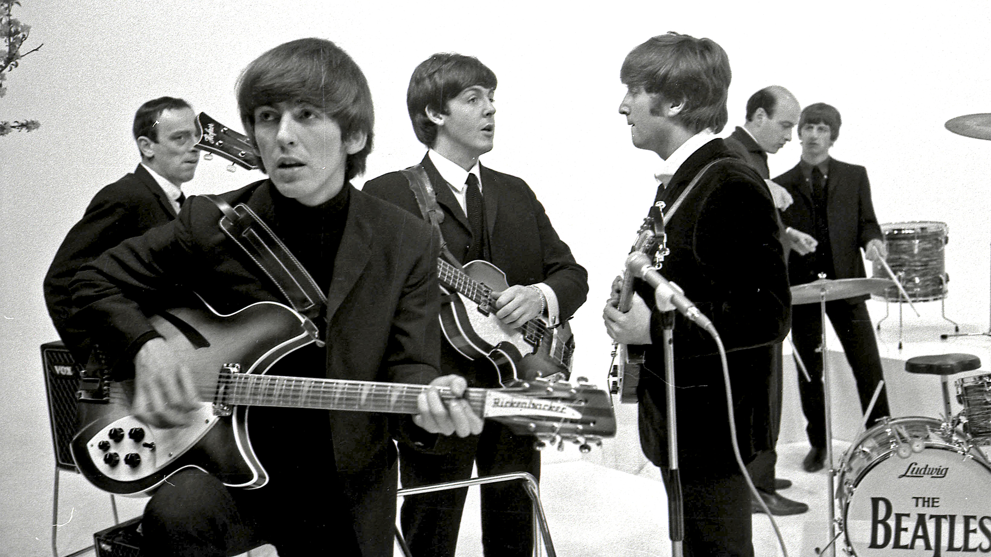 The Beatles on the set of A Hard Day's Night. L-R: John Junkin, George Harrison (playing Rickenbacker 360/12 guitar), Paul McCartney, John Lennon, Richard Lester (director), Ringo Starr during the filming of "A Hard Day's Night" at Scala Theatre