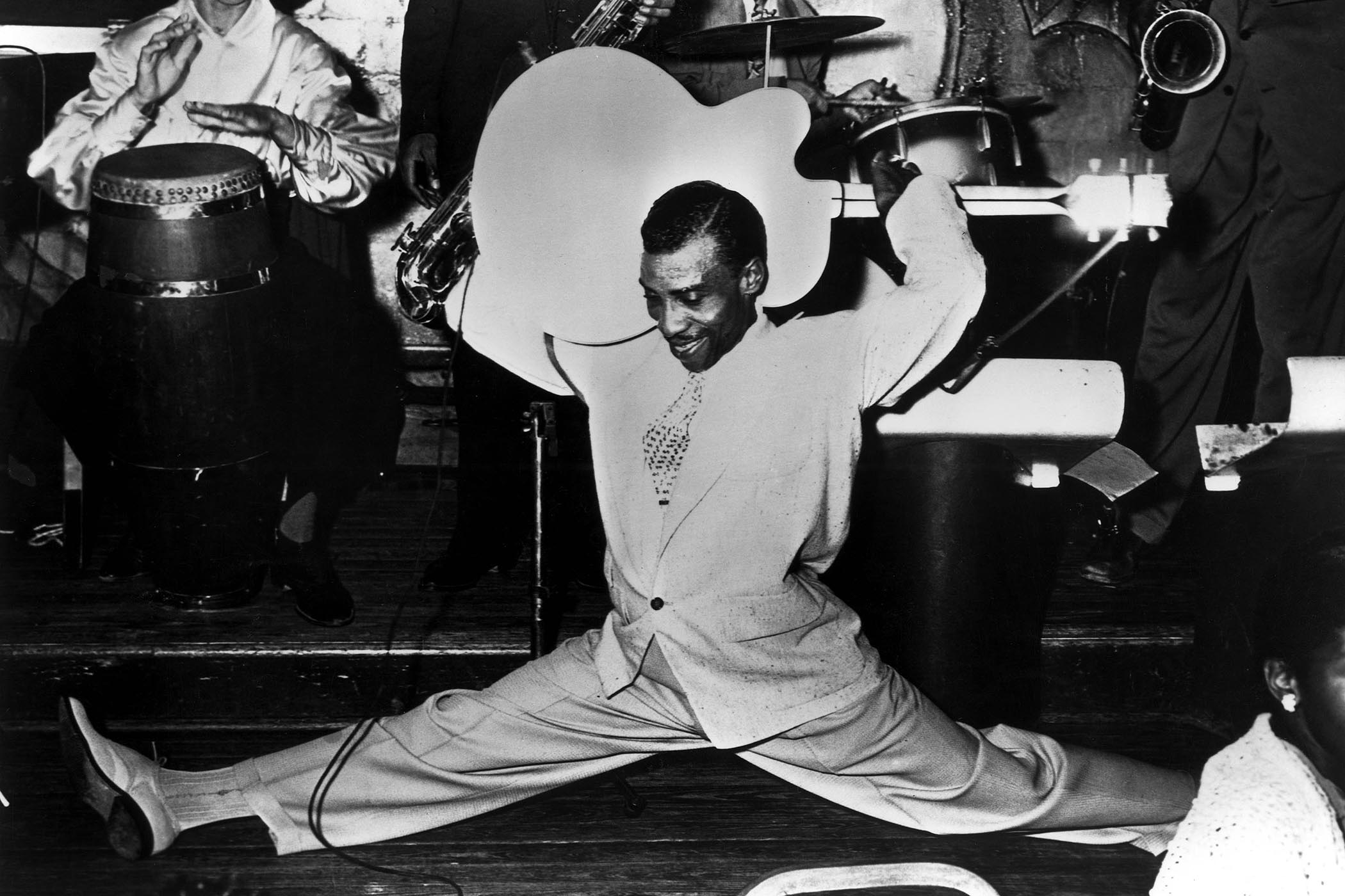 T-Bone Walker does the splits while playing the guitar behind his head in this black-and-white image from the '50s.