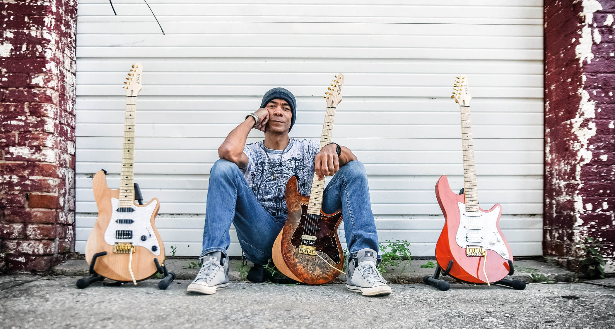 Greg Howe sits in front of his garage door with three of his S-style electric guitars