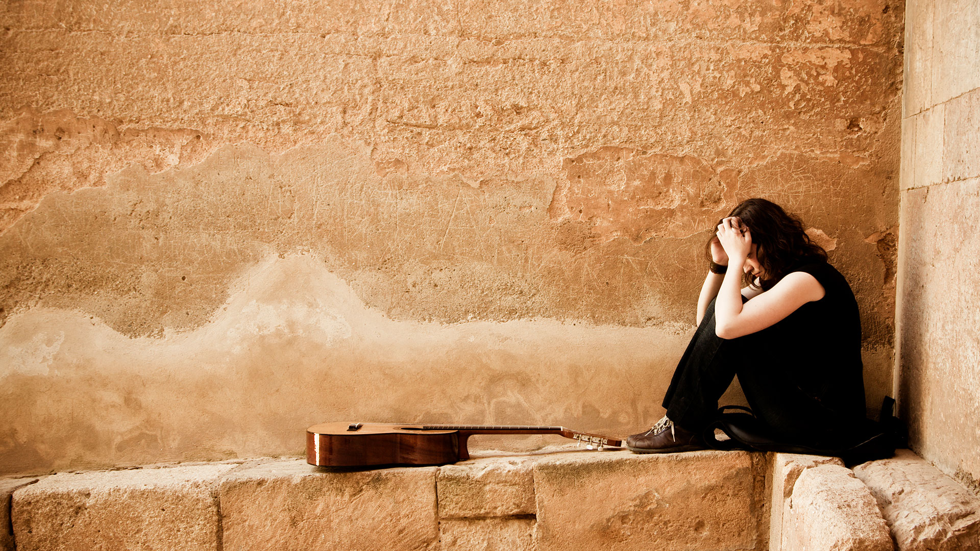 A musician sits with her head in her hands and her guitar lying next to her, in front of a stone wall
