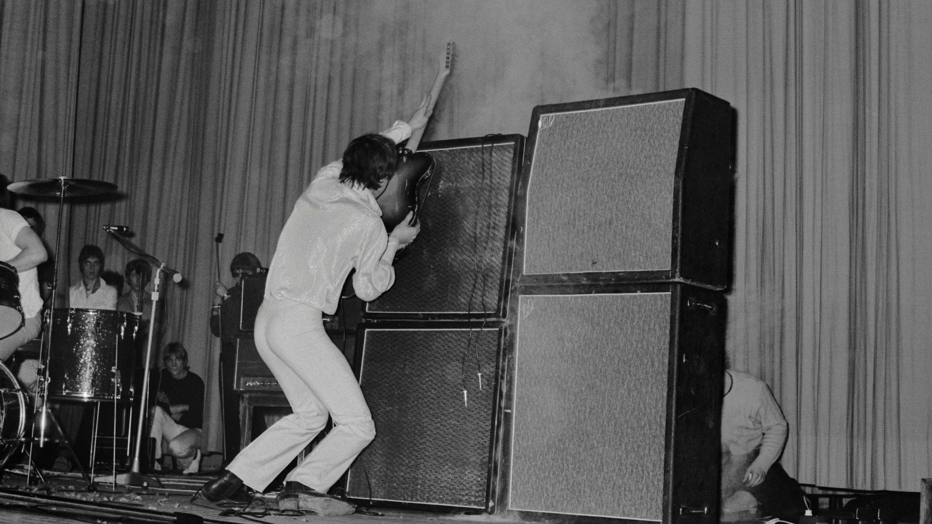 Pete Townshend hold his guitar up against a stack of guitar cabinets