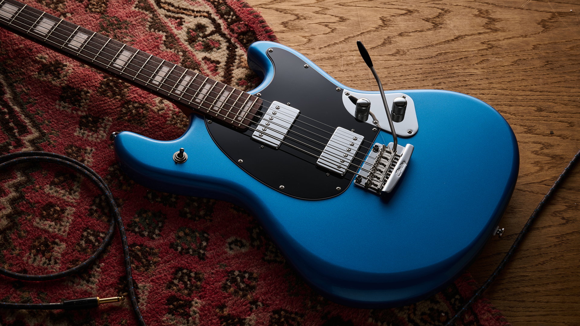 A Sterling by Music Man StingRay Baritone guitar lying on a rug and wooden floor with a guitar cable coiled nearby
