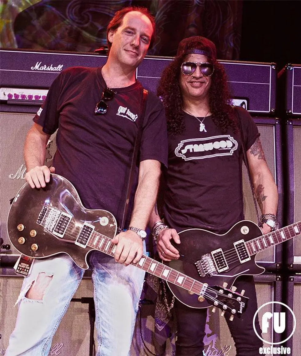 Two guitarists stand together with electric guitars in front of amplifiers, smiling.